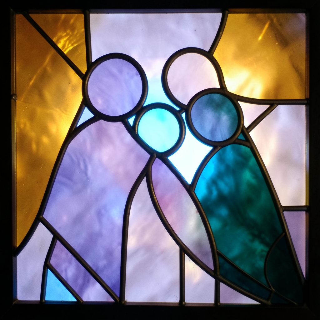 A stained-glass composition with bold leaded segments in warm amber, soft lavender, and deep teal, forming an abstract family silhouette against a glowing background.