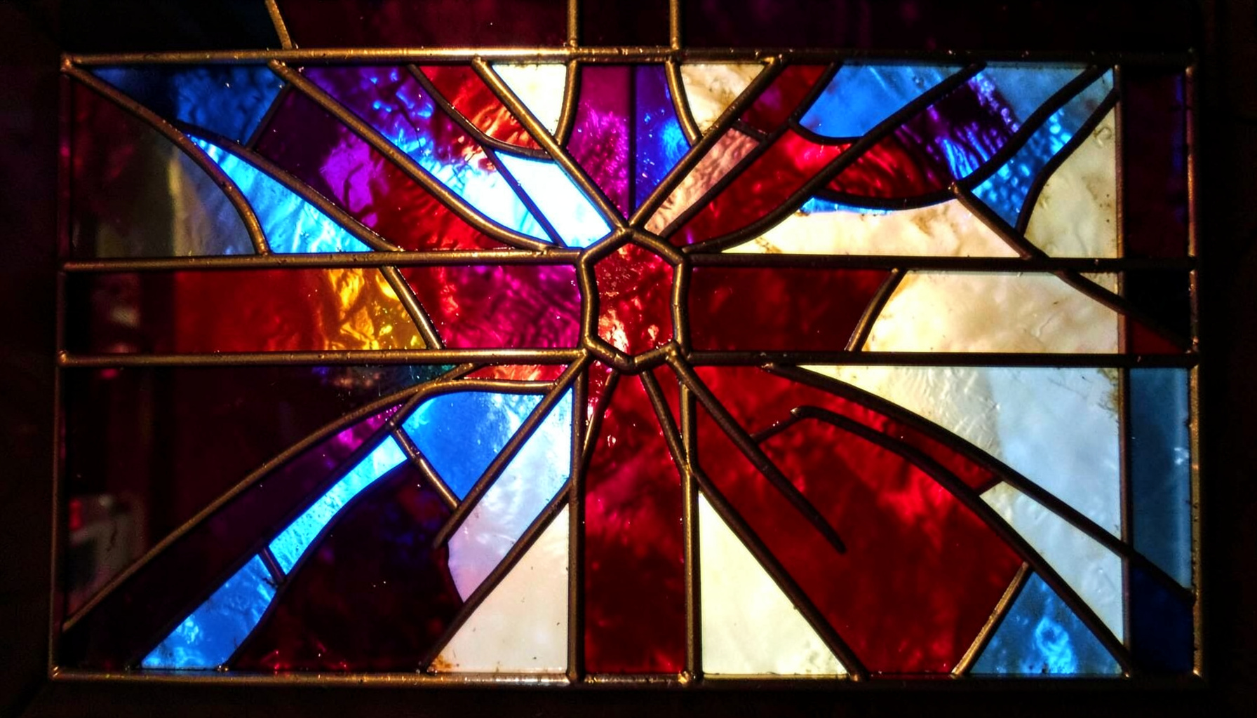A stained-glass piece in rich cranberry, gold, and ivory with tight leaded segments forming a festive, gift-like pattern.